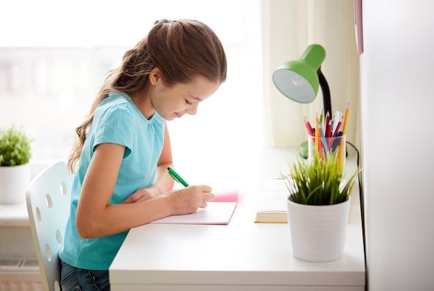 people, children, education and learning concept - happy girl writing to notebook and doing homework at home