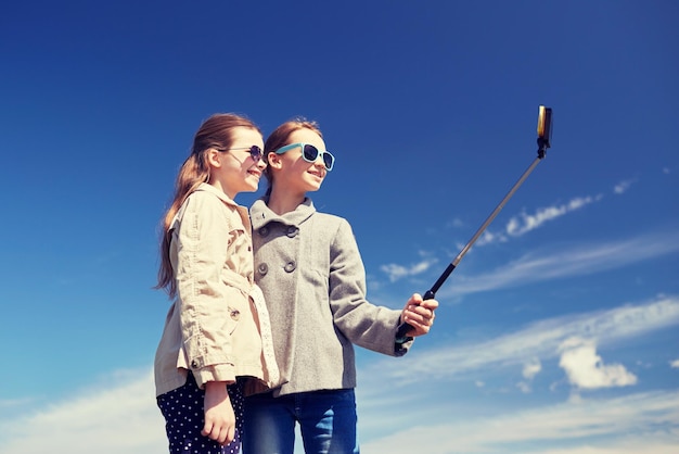 人、子供、友達、友達のコンセプト – 幸せな女の子が屋外で自撮り棒でスマートフォンで写真を撮る