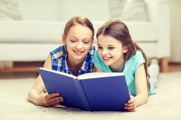 people, children, friends, literature and friendship concept - two happy girls lying on floor and reading book at home