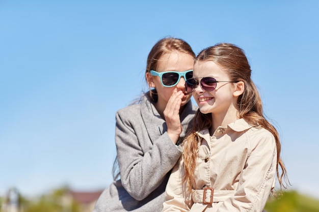 люди, дети и концепция дружбы - счастливая маленькая девочка в солнечных очках шепчет свой секрет друзьям на ухо или сплетничает на улице