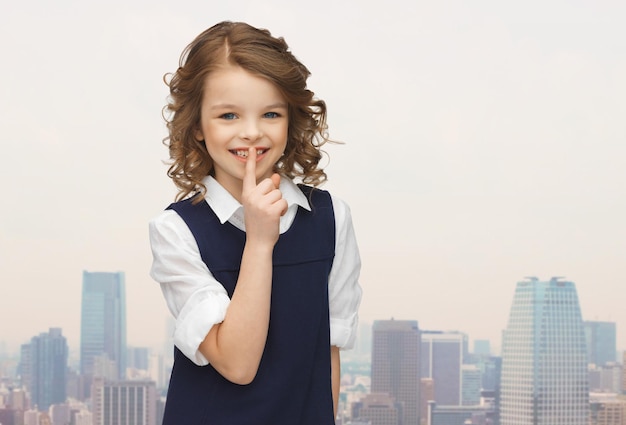 people, children, secrecy and mystery concept - happy girl showing hush gesture over city background