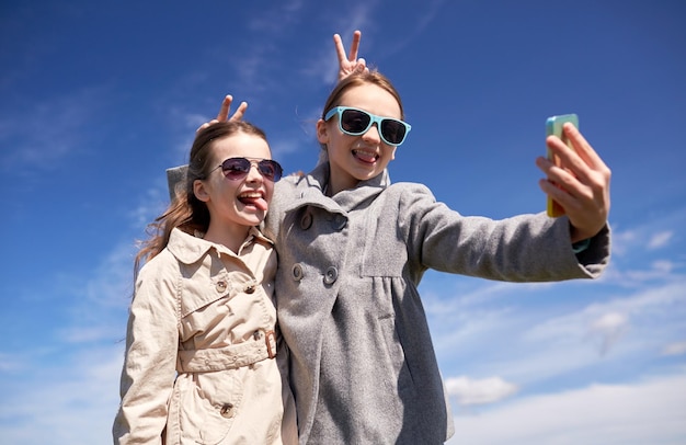 people, children, technology, friends and friendship concept - happy girls with smartphone taking selfie and making funny faces outdoors