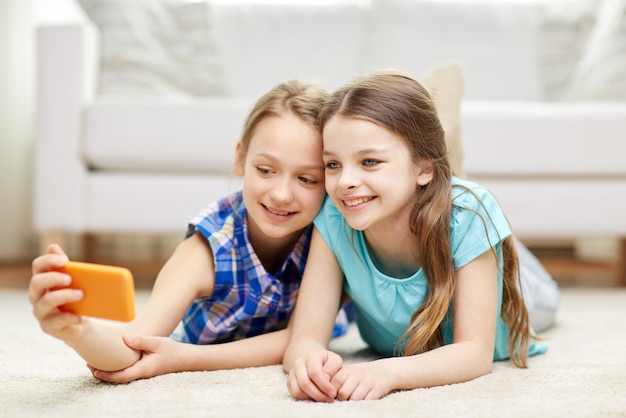 people, children, technology, friends and friendship concept - happy little girls lying on floor and taking selfie with smartphone at home