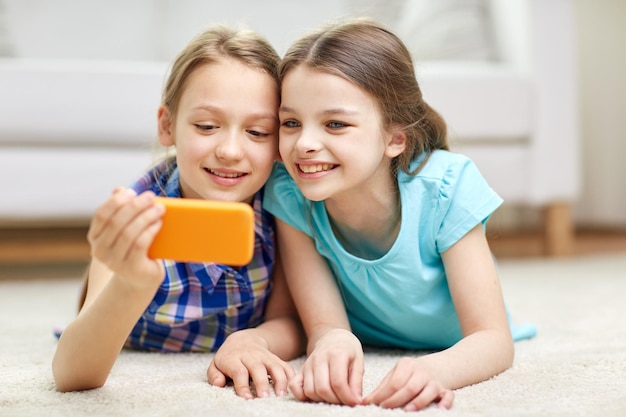 人、子供、技術、友人、友情のコンセプト – 幸せな小さな女の子が床に横たわり、自宅でスマートフォンを使って自撮りをする