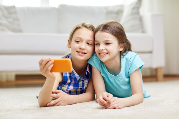 人、子供、技術、友人、友情のコンセプト – 幸せな小さな女の子が床に横たわり、自宅でスマートフォンを使って自撮りをする