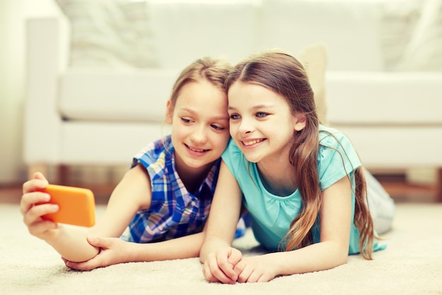 people, children, technology, friends and friendship concept - happy little girls lying on floor and taking selfie with smartphone at home