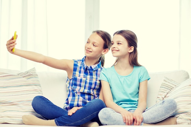人、子供、技術、友達、友情のコンセプト – ソファに座り、自宅でスマートフォンを使って自撮りをする幸せな女の子