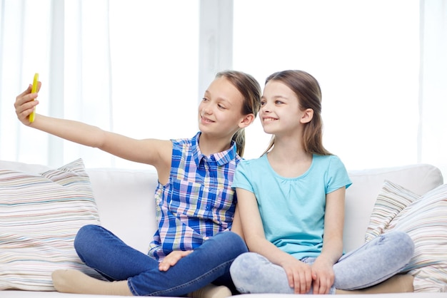人、子供、技術、友達、友情のコンセプト – ソファに座り、自宅でスマートフォンを使って自撮りをする幸せな女の子