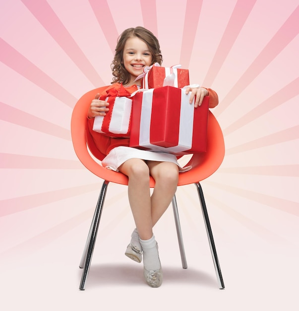 people, christmas, holidays, presents and childhood concept - happy little girl with gift boxes sitting on chair over pink burst rays background