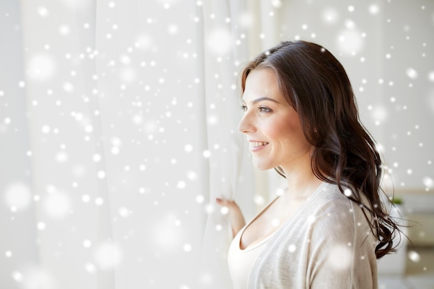 people, christmas, winter and leisure concept - happy young woman looking through window at home over snow