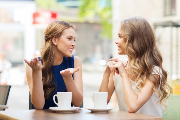 people, communication and friendship concept - smiling young women drinking coffee or tea and talking at outdoor cafe