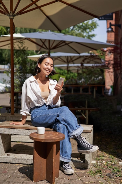 Photo people and communication stylish asian girl sits in outdoor cafe with cup of coffee and smartphone u