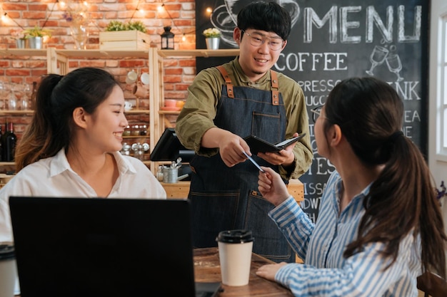 people consumerism lifestyle and friendship concept. smiling young male waiter in apron taking credit card from customer hand. lady coworkers paying for coffee at cafe bar while working on laptop