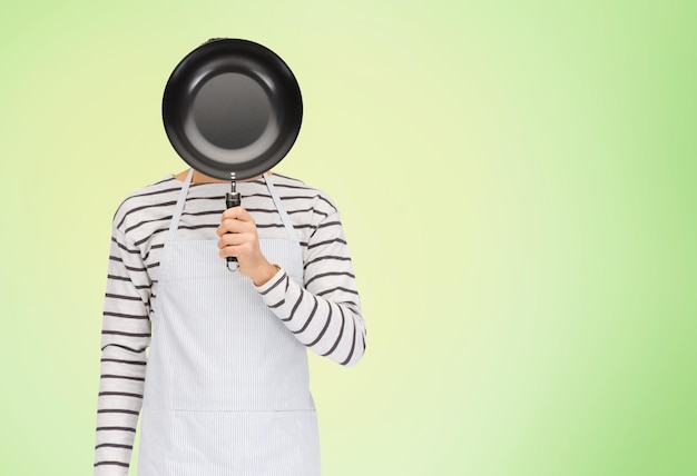 Photo people, cooking, culinary and identity concept - man or cook in apron hiding his face behind frying pan over green natural background