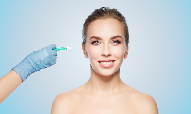 people, cosmetology, plastic surgery and beauty concept - beautiful young woman face and beautician hand in glove with syringe making injection over blue background