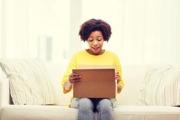 people, delivery, shipping and postal service concept - happy african american young woman opening cardboard box or parcel at home