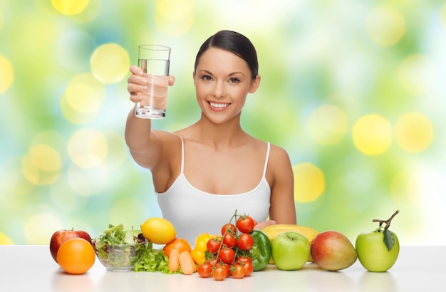 people, diet, healthy eating and food concept - happy woman with glass of water, fruits and vegetables on table over green summer lights background