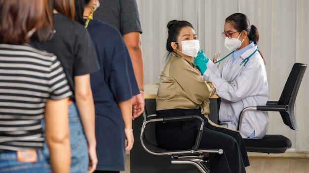 의사가 얼굴 마스크를 쓴 노인 여성에게 독감이나 폐렴 주사를 놓는 동안 다양한 연령대의 사람들이 연속으로 서서 백신 주사를 기다리고 있습니다. 코비드-19 또는 코로나바이러스 예방 접종 개념.