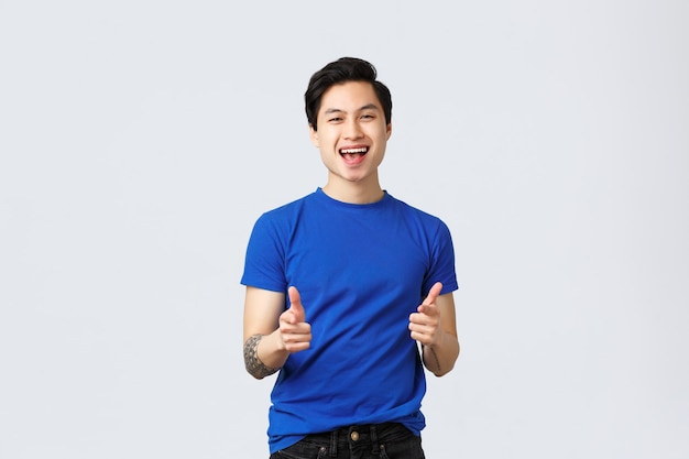 People different emotions, lifestyle and casual concept. Cheerful upbeat asian man in blue t-shirt, pointing fingers up, gotcha gesture, congratulate person with big win, grey background