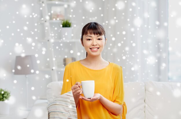 people, drinks and leisure concept - happy asian woman sitting on sofa and drinking tea from cup or mug at home over snow