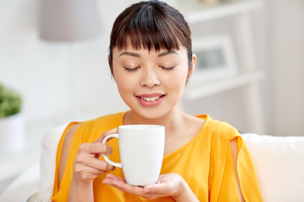 people, drinks and leisure concept - happy asian woman sitting on sofa and drinking tea from cup or mug at home