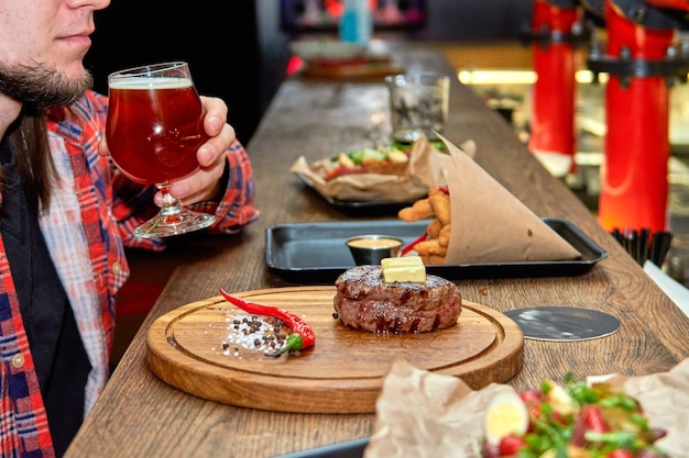 People eating at fast food spending time together in cafe, beer pub. Entrecote Beef Grilled steak meat on wooden cutting boardon with  pepper and salt with bear at the bar counter.