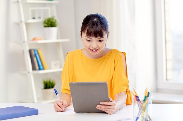 people, education, high school and learning concept - happy asian young woman student with tablet pc computer, book and notepad writing at home