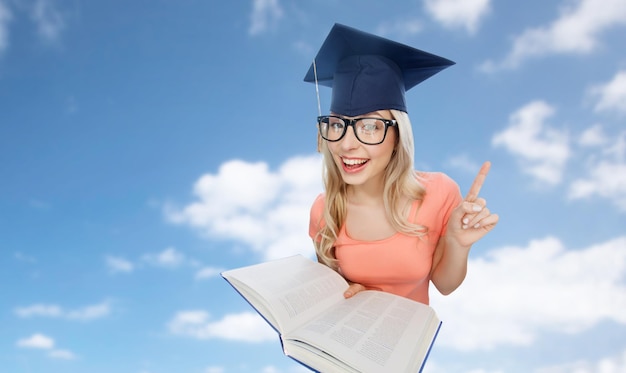 people, education, knowledge and graduation concept - smiling young student woman in mortarboard and eyeglasses with encyclopedia book pointing finger up over blue sky and clouds background