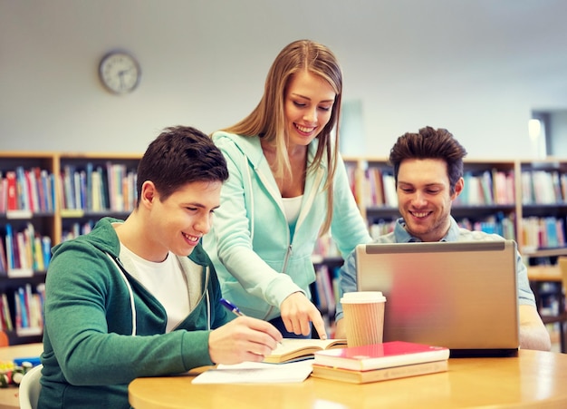 people, education, technology and school concept - happy students with laptop computer and books in library