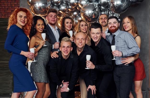 People in elegant clothes with drinks in hands standing together and celebrating New Year holidays in christmas decorated room.