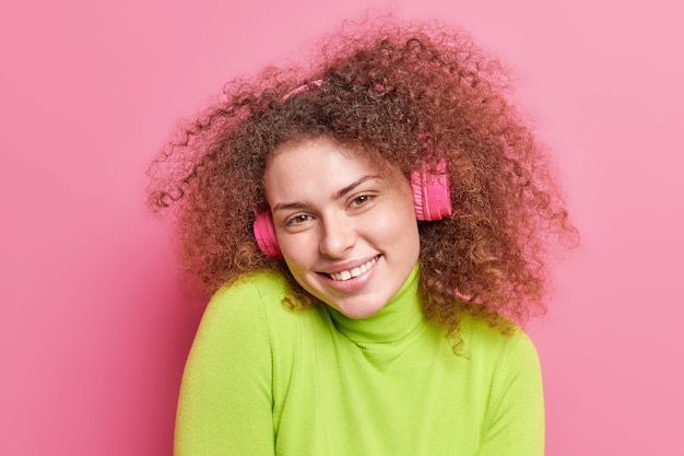 People emotions hobby concept. Positive cury haired teenage girl smiles gladfully expresses joy wears wireless headphones on ears enjoys listening music wears green jumper isolated on pink wall