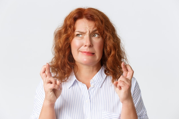 Photo people, emotions and lifestyle concept. close-up of funny worried middle-aged redhead woman grimacing while cross fingers good luck, making wish and feeling nervous, white wall.