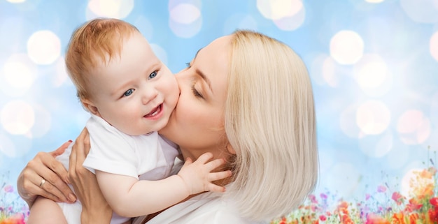 people, family, motherhood and children concept - happy mother hugging adorable baby over blue lights and poppy field background