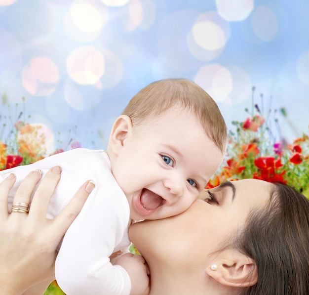 people, family, motherhood and children concept - happy mother hugging adorable baby over blue lights and poppy field background