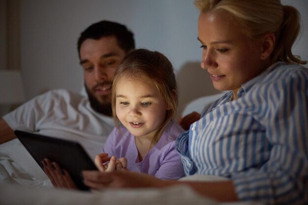 people, family and technology concept - happy mother, father and little girl with tablet pc computer in bed at night home