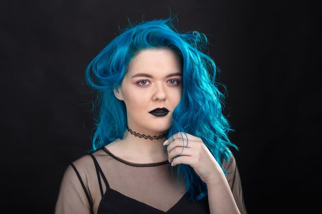 Люди и концепция моды - молодая и привлекательная женщина с черной помадой и синими волосами, позирующая над черной стеной.