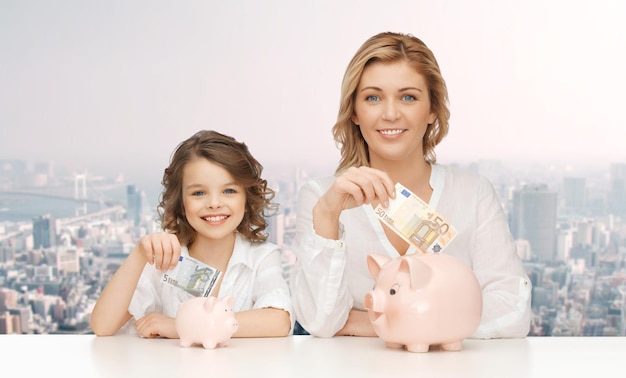 people, finances, family budget and savings concept - happy mother and daughter with piggy banks and paper money over city background