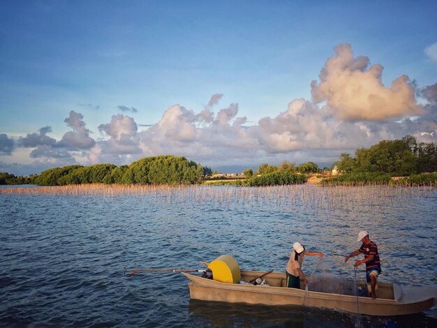 People fishing while standing in boat of lake against sky