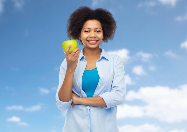 人、食べ物、健康的な食事、歯科医療のコンセプト – 青い空と雲の背景に緑のリンゴを持つ幸せなアフリカ系アメリカ人の若い女性
