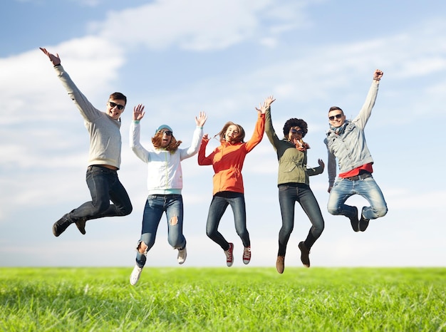 people, freedom, happiness and teenage concept - group of happy friends in sunglasses jumping high over blue sky and grass background