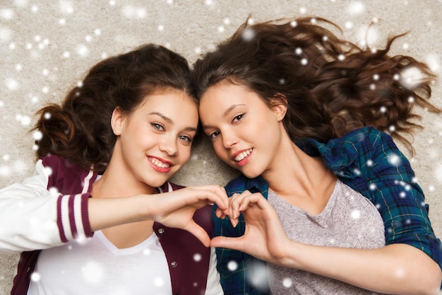 Photo people, friends, winter, christmas and friendship concept - happy smiling pretty teenage girls lying on floor showing heart sign over snow