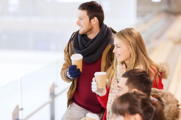 Persone, amicizia, bevande calde e concetto di svago - amici felici che bevono da tazze di caffè di carta sulla pista di pattinaggio