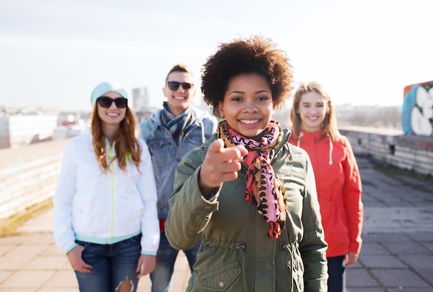 people, friendship and international concept - happy african american young woman or teenage girl pointing finger to you in front of her friends on city street