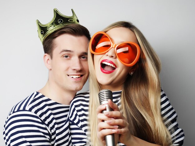 Concetto di persone, amicizia, amore e tempo libero - bella giovane coppia amorosa con microfono. grandi occhiali e corona arancioni. pronto per la festa.