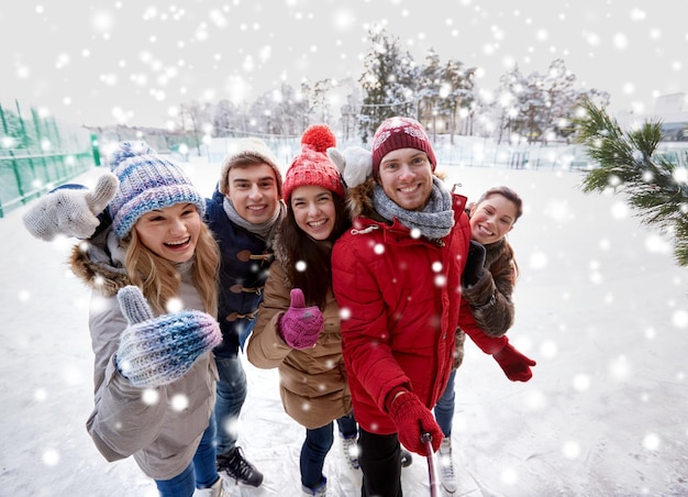 Concetto di persone, amicizia, tecnologia e tempo libero - amici felici che scattano foto con il selfie stick dello smartphone e mostrano i pollici in su sulla pista di pattinaggio su ghiaccio all'aperto