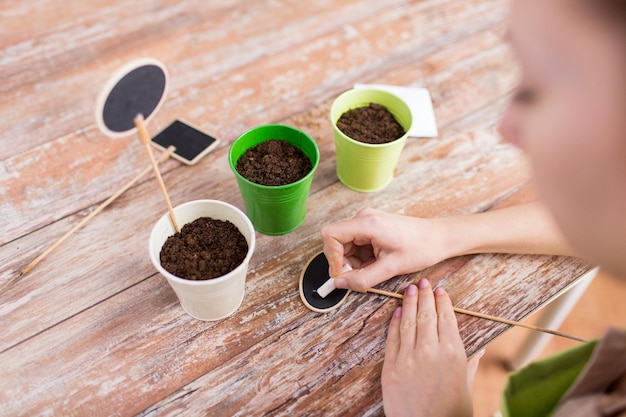 people, gardening, seeding and profession concept - close up of woman writing name on garden sign over pots with soil and seeds