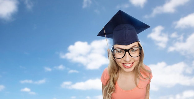 people, graduation and education concept - smiling young student woman in mortarboard and eyeglasses over blue sky and clouds background