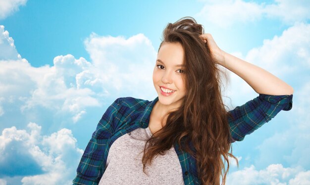 People, hair care, style and teens concept - happy smiling pretty teenage girl touching her head over blue sky and clouds background