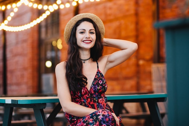 Photo people happiness and fashion concept attractive female with wellshaped eyebrows dark eyes and red rouged lips wearing straw hat and dress sitting at cafeteria admiring good atmosphere