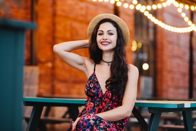 People happiness and fashion concept Attractive female with wellshaped eyebrows dark eyes and red rouged lips wearing straw hat and dress sitting at cafeteria admiring good atmosphere